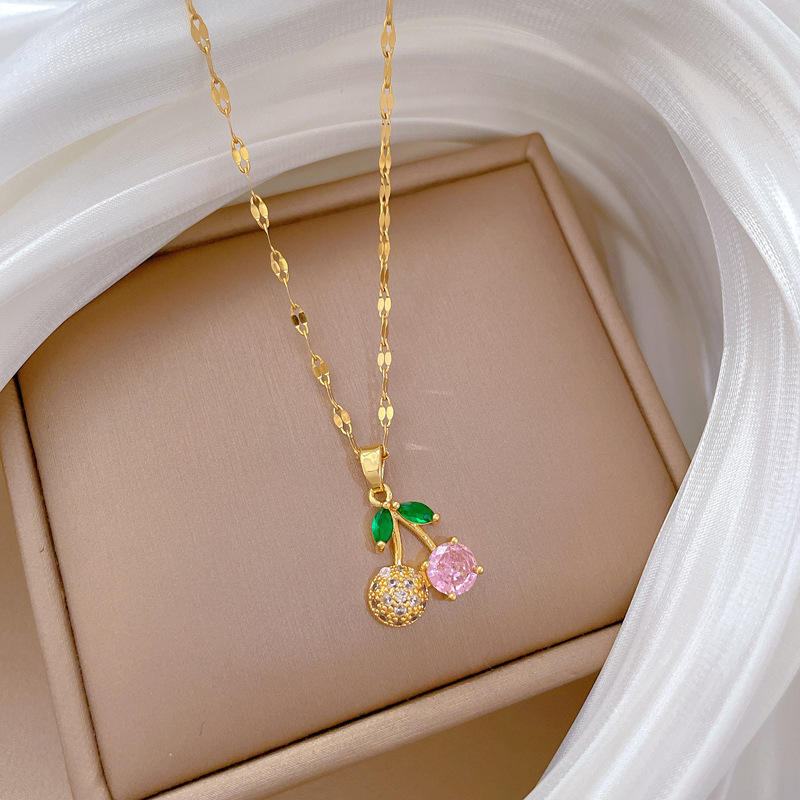 Close-up of gold-tone cherry pendant necklace featuring pink and clear crystals and green leaf details on beige background