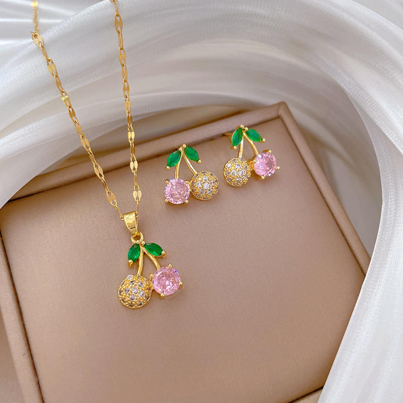Gold-tone cherry pendant necklace and earrings set displayed on beige background with white fabric drape